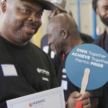 A male employee smiles and holds up a sign saying Own Together, Achieve Together, Marmic Pride. 